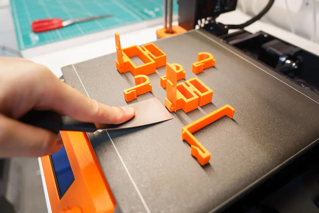 Une main décolle à la spatule des pièces orange tout juste imprimées sur le plateau d’une imprimante 3D