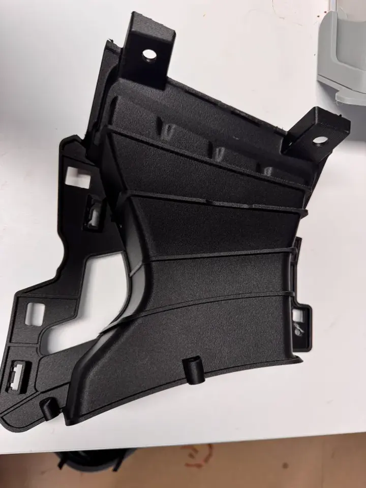 Pièce technique noire imprimée en 3D, aux formes complexes avec pattes de fixation, posée sur une table claire