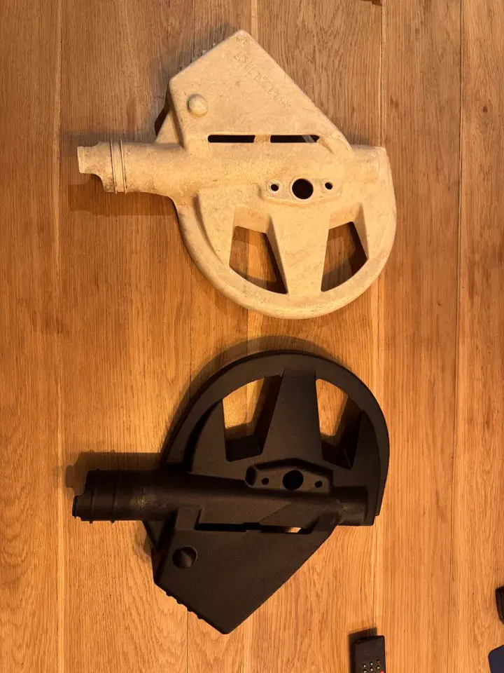 Une pièce d’origine claire et cassée posée à côté de sa copie noire imprimée en 3D, sur un plan de travail en bois
